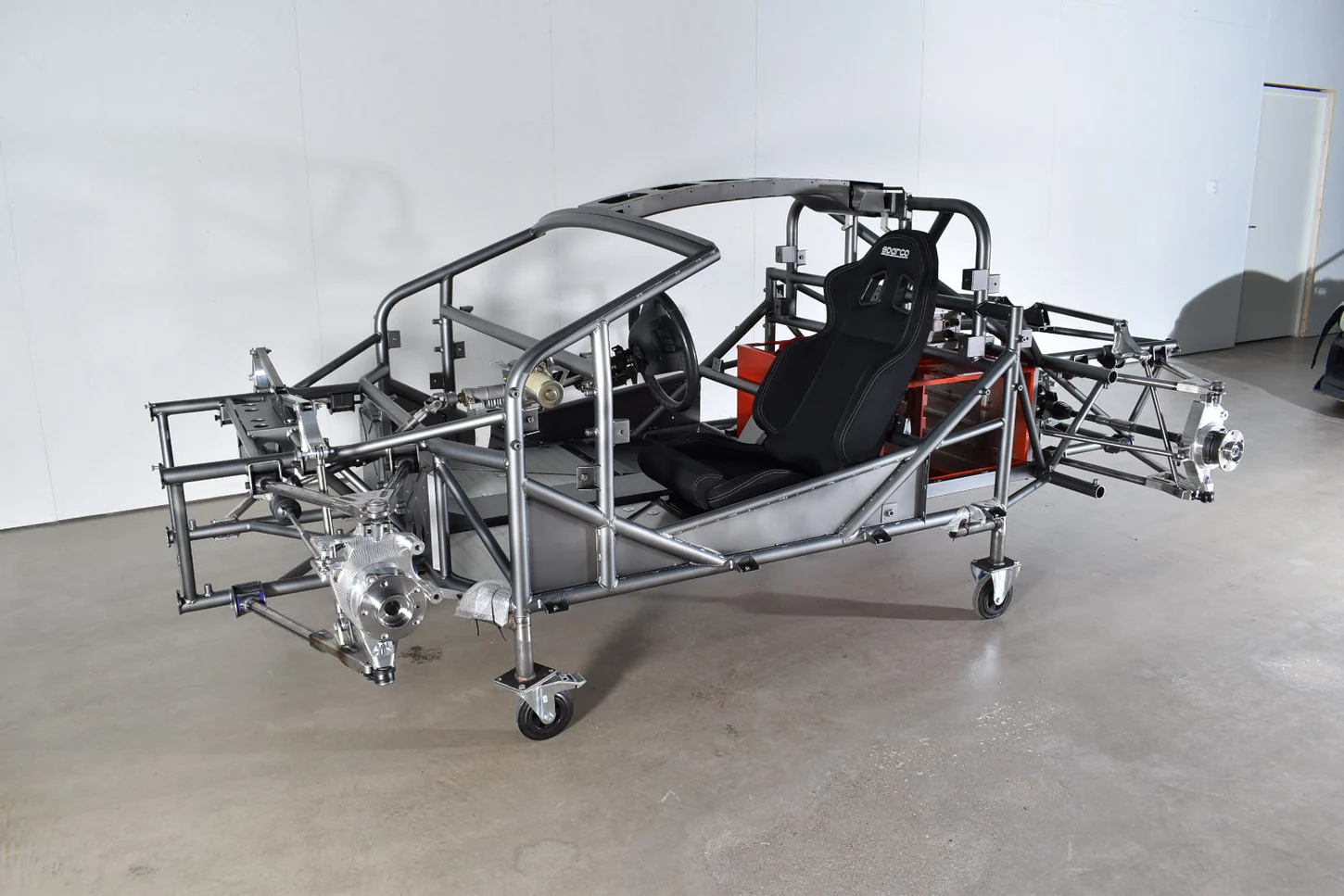 Unbodied welded tubular vehicle chassis with driver's seat and exposed suspension uprights on workshop castors