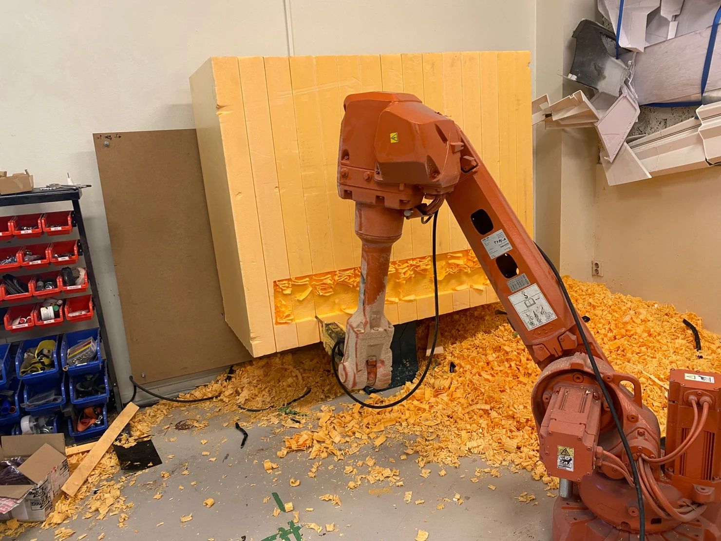 Orange industrial robot milling a large foam block amid deep piles of orange shavings in a workshop